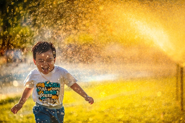 A small toddler smiles as he runs through the sprinklers in a yard on a hot summer's day. Photo by MI PHAM on Unsplash.