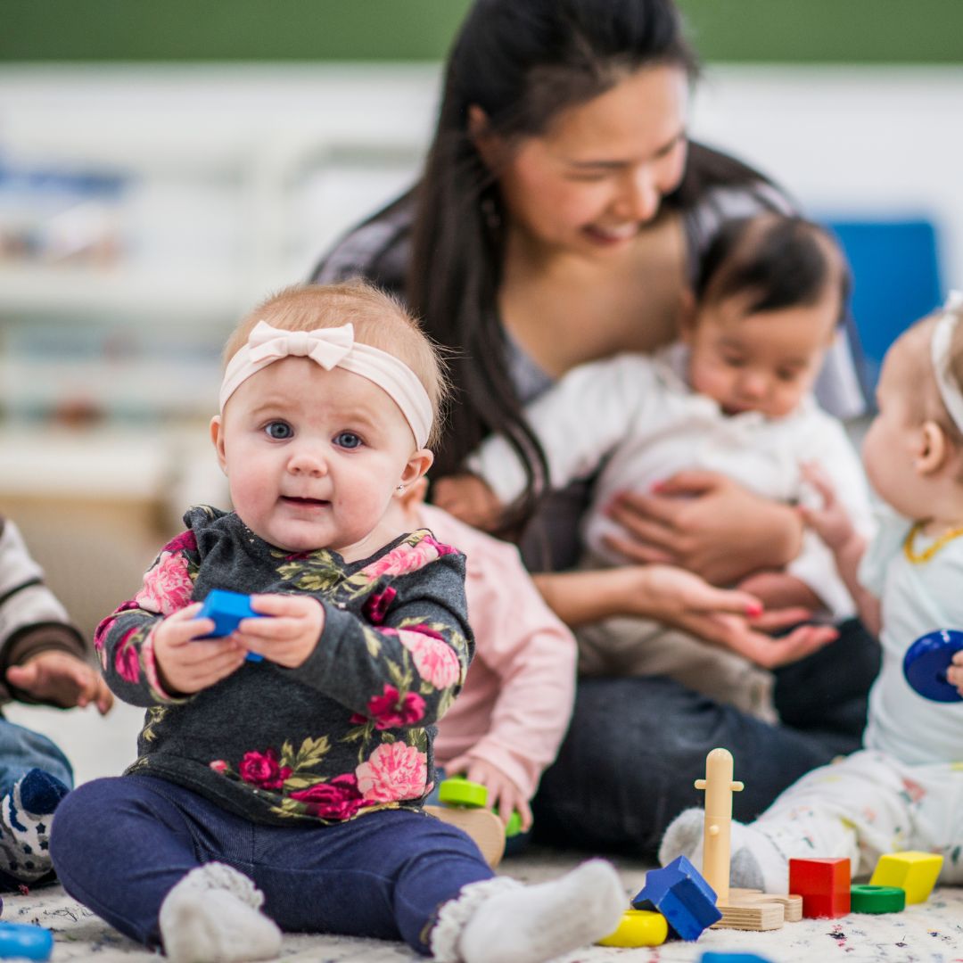 Infant smiling at camera, other babies and daycare teacher in background