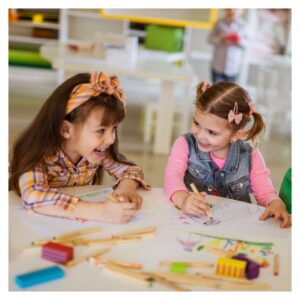 girls sit at table drawing pictures and laughing together 